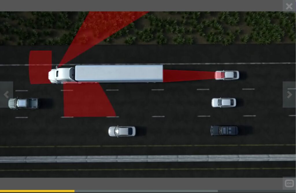 Screen shot of a training video for truck drivers. Shows an overhead view of a white 18-wheeler and several other cars on a 3-lane highway moving right to left. There are red areas projecting from all sides of the truck indicating blind spots.